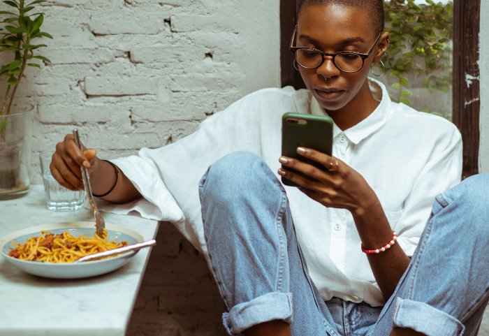 Femme noire qui mange un spaguetti en regardant son téléphone cellulaire.